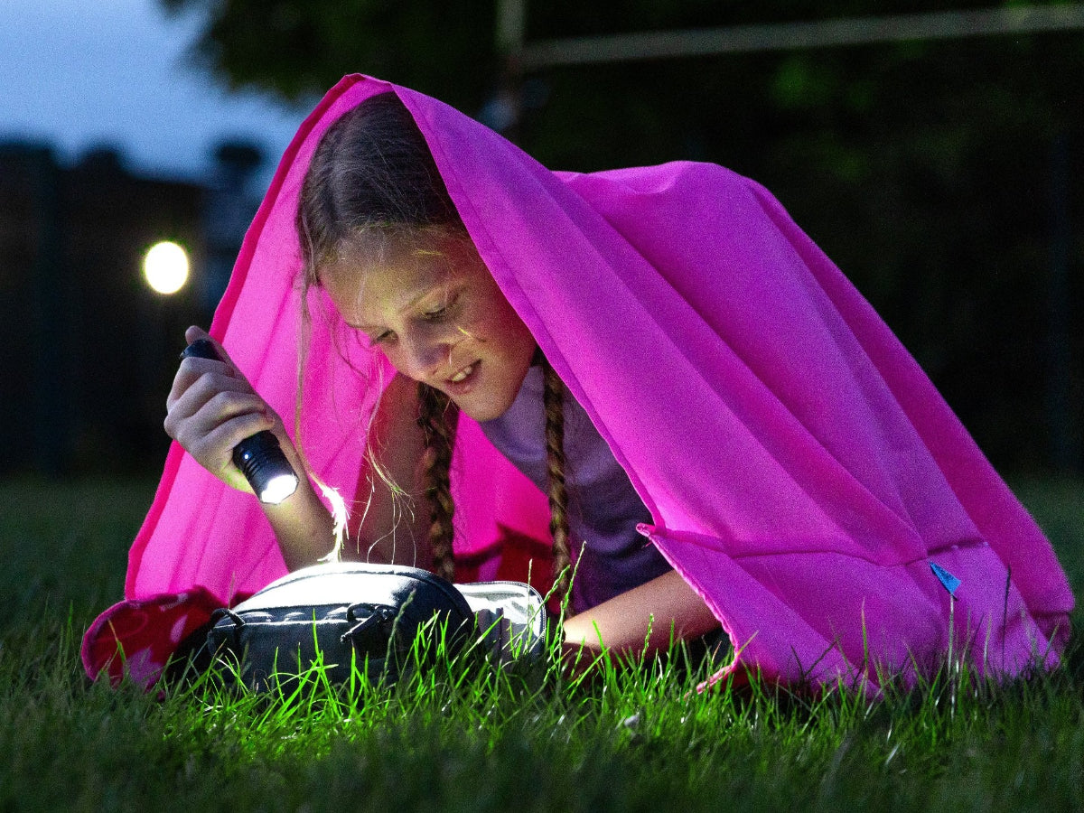 A young girl laying down inside a pink GIGSAK Kids outdoor waterproof blanket, the only outdoor waterproof blanket you can sit inside. She is reading, but it is dark so she is using a flashlight to read.