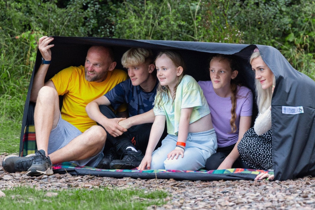 A large family sat inside a GIGSAK Giant outdoor waterproof blanket, the only outdoor waterproof blanket you can sit inside. It features a sleek red, orange, yellow, and green tartan with a black waterproof design. GIGSAK features a unique, hidden, cosy shelter to protect against the wind and rain. Simply turn GIGSAK inside out to stay warm and dry. GIGSAK is perfect for family picnics, BBQs, the beach, festivals, open-air cinema, fireworks, camping, and more.