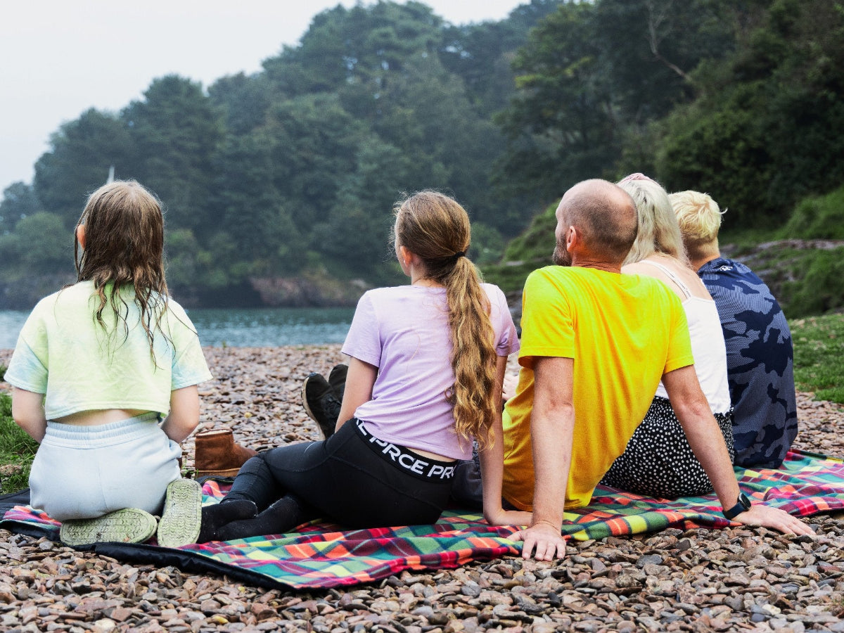 A family of five sat on a GIGSAK Giant outdoor waterproof blanket, the only outdoor waterproof blanket you can sit inside. It features a sleek red, orange, yellow, and green tartan with a black waterproof design. GIGSAK features a unique, hidden, cosy shelter to protect against the wind and rain. Simply turn GIGSAK inside out to stay warm and dry. GIGSAK is perfect for family picnics, BBQs, the beach, festivals, open-air cinema, fireworks, camping, and more.