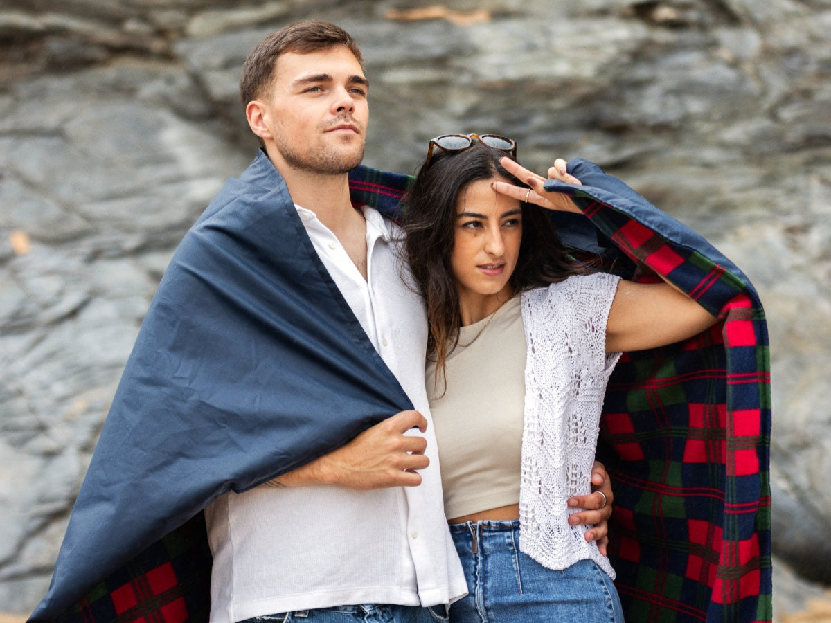 A young couple wrapped in a GIGSAK outdoor waterproof blanket, the only outdoor waterproof blanket you can sit inside. Features a sleek red, blue, and green coloured tartan with navy blue waterproof design. GIGSAK features a unique, hidden, cosy shelter to protect against the wind and rain. Simply turn GIGSAK inside out to stay warm and dry. GIGSAK is perfect for family picnics, BBQs, the beach, festivals, open-air cinema, fireworks, camping, and more.