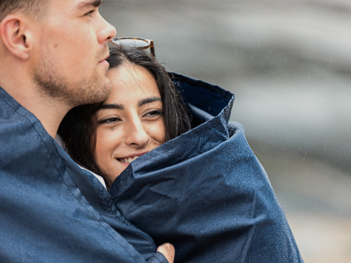 A young couple wrapped in a GIGSAK outdoor waterproof blanket, the only outdoor waterproof blanket you can sit inside. Features a sleek red, blue, and green coloured tartan with navy blue waterproof design. GIGSAK features a unique, hidden, cosy shelter to protect against the wind and rain. Simply turn GIGSAK inside out to stay warm and dry. GIGSAK is perfect for family picnics, BBQs, the beach, festivals, open-air cinema, fireworks, camping, and more.