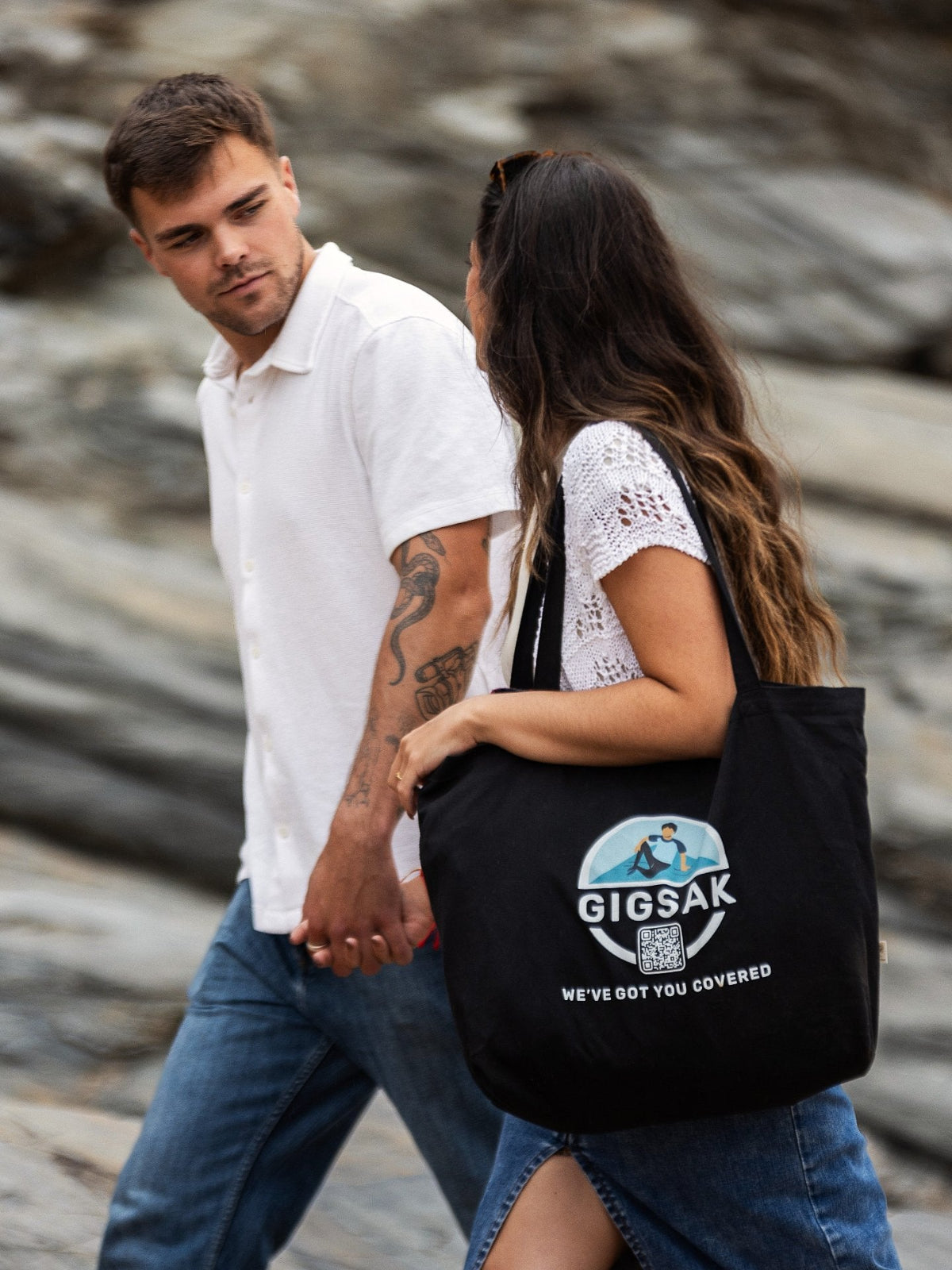 A young couple walking with a GIGSAK large tote bag on the girl's shoulder. The GIGSAK outdoor waterproof blanket is the only outdoor waterproof blanket you can sit inside. Features a sleek red, blue, and green coloured tartan with navy blue waterproof design. GIGSAK features a unique, hidden, cosy shelter to protect against the wind and rain. Simply turn GIGSAK inside out to stay warm and dry. GIGSAK is perfect for family picnics, BBQs, the beach, festivals, open-air cinema, fireworks, camping, and more.