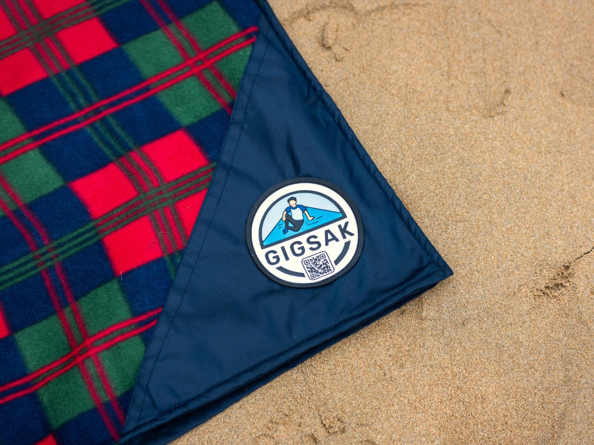 A GIGSAK outdoor waterproof blanket, the only outdoor waterproof blanket you can sit inside. Features a sleek red, blue, and green coloured tartan with navy blue waterproof design. GIGSAK features a unique, hidden, cosy shelter to protect against the wind and rain. Simply turn GIGSAK inside out to stay warm and dry. GIGSAK is perfect for family picnics, BBQs, the beach, festivals, open-air cinema, fireworks, camping, and more.